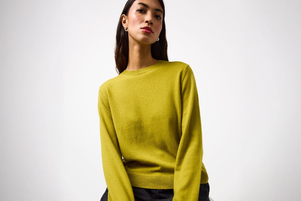 Person wearing a bright yellow long-sleeve crew-neck sweater paired with dark grey jeans, seated on a light-colored surface.