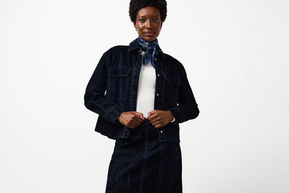 Person wearing a dark denim jacket and matching mini skirt, styled with a white top, blue patterned neck scarf, brown socks, and black loafers.