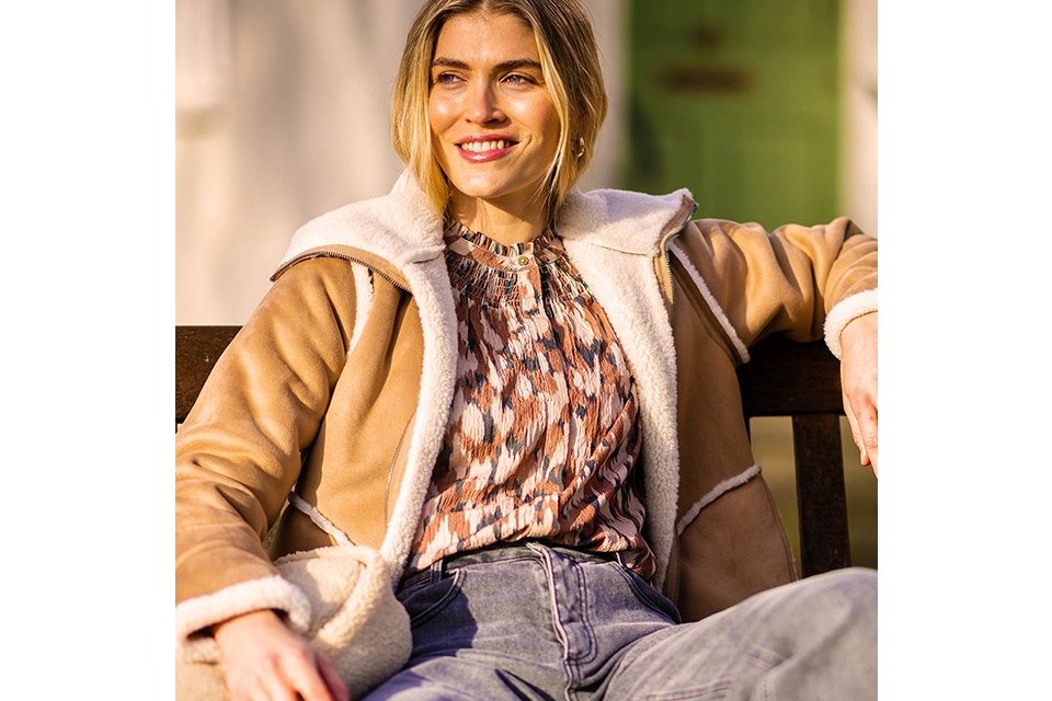Person sitting on a wooden bench wearing a tan shearling-lined jacket over a patterned blouse, paired with high-waisted light blue jeans and holding a cream textured bag.