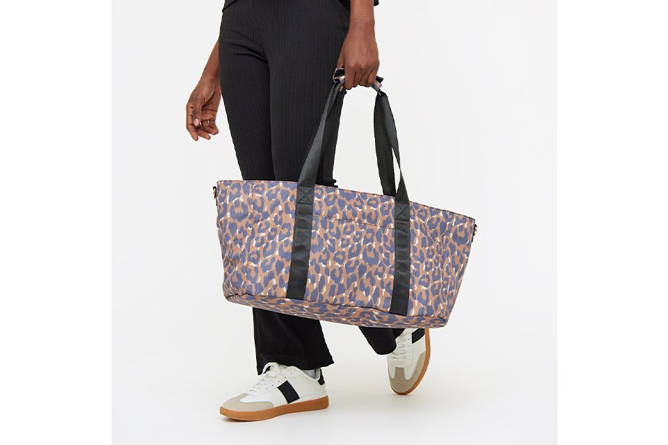 Person wearing a black textured top and matching pants, holding a large rectangular baby changing bag with a blue and brown leopard print design and black shoulder straps. The person is also wearing white sneakers with black and tan accents.