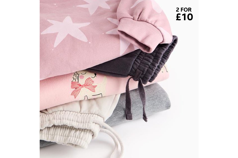 Stack of pastel joggers and sweatshirts, including pink, grey, and star‑print pieces.