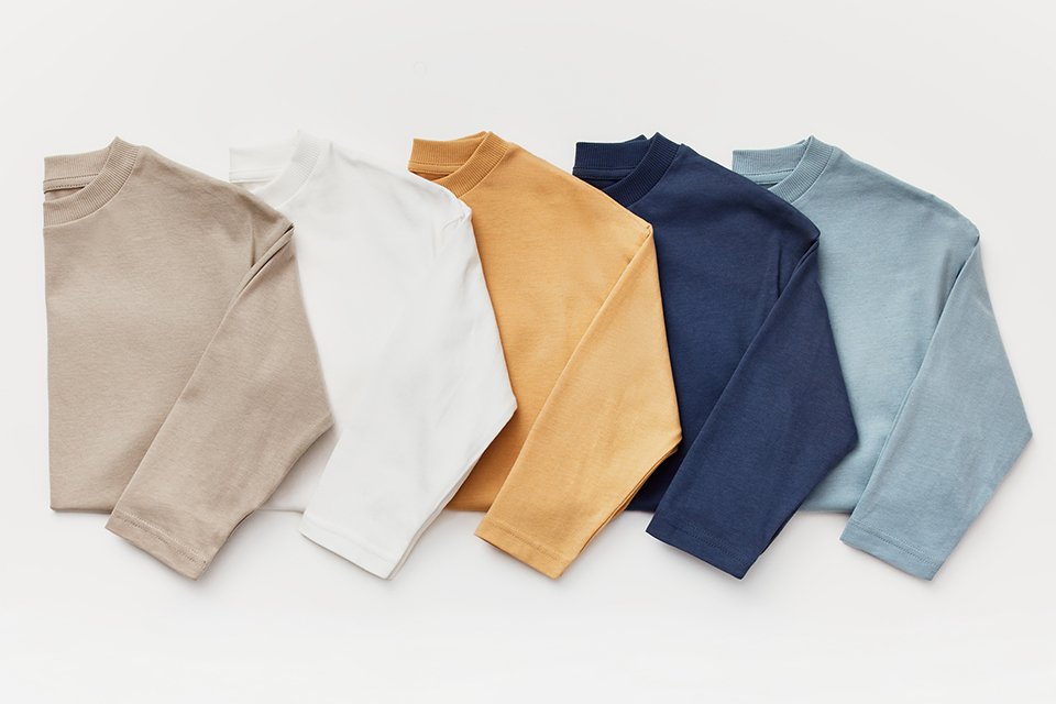 Five long-sleeve tops in neutral and muted tones—beige, white, mustard, navy, and light blue—arranged in a row.