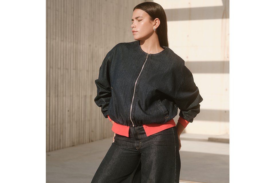 Person wearing a dark denim zip-up jacket with a bright red waistband detail, paired with matching wide-leg dark denim jeans and black shoes, standing in an outdoor setting with a light concrete background. 