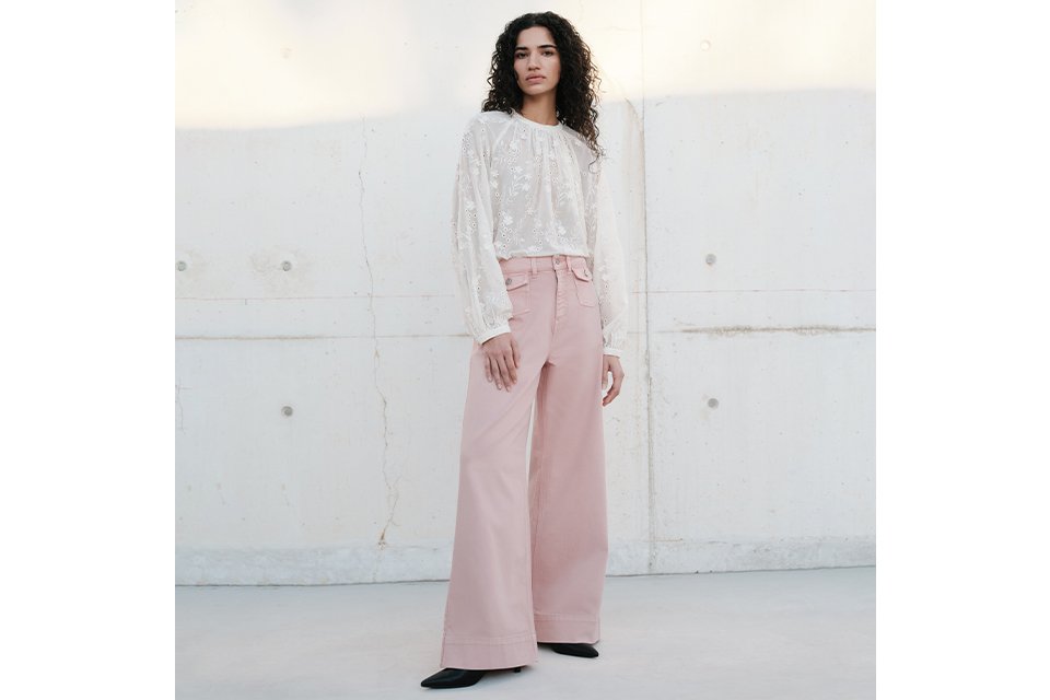 Person standing against a light concrete wall wearing a long-sleeved white lace blouse paired with high-waisted, wide-leg pink trousers and black pointed shoes. 