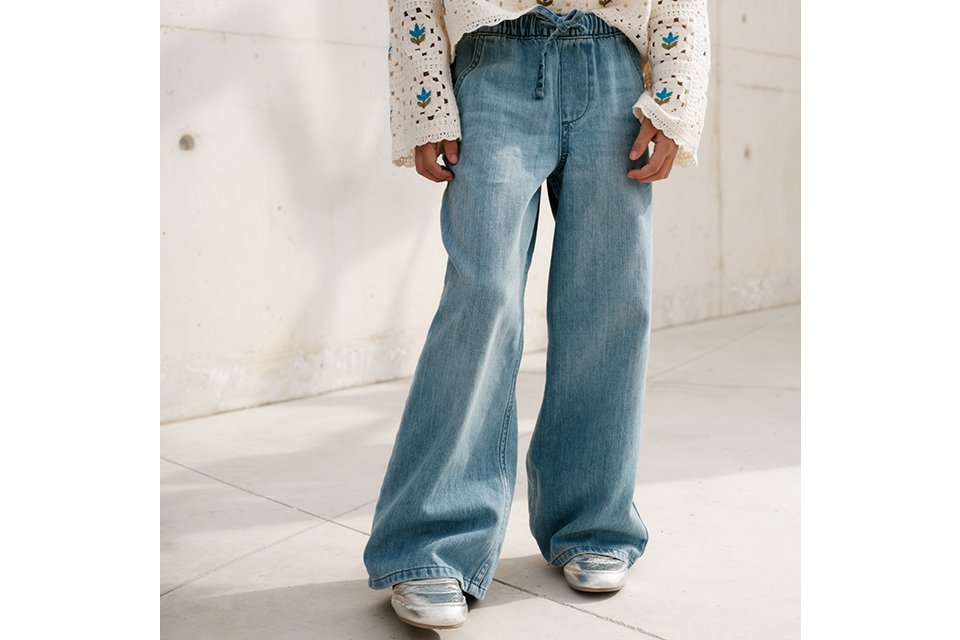 Child wearing light blue wide-leg jeans paired with a cream crochet-style sweater featuring a blue and green floral pattern, and silver shoes, standing against a light concrete wall.