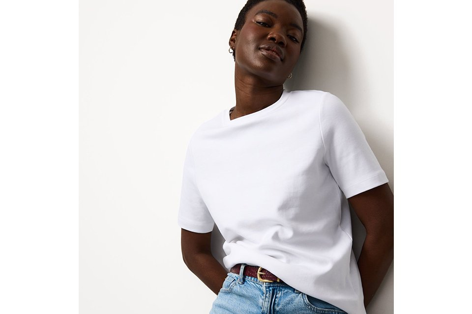 Person wearing a plain white short-sleeve T-shirt tucked into light blue high-waisted jeans with a brown belt, leaning casually against a white wall.