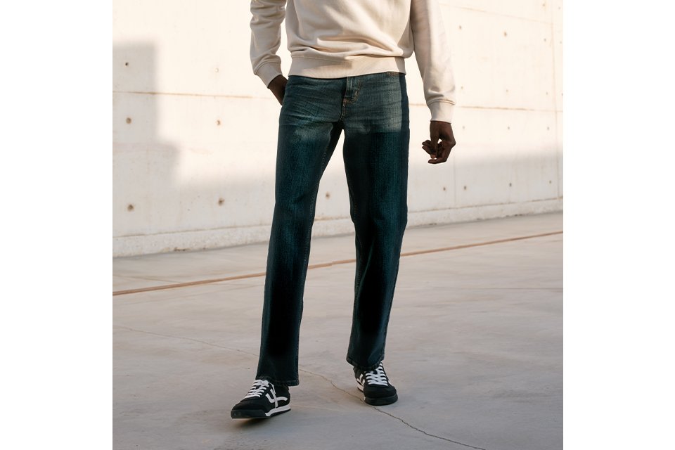 Person wearing a cream long-sleeve sweatshirt paired with dark blue straight-leg jeans and black-and-white sneakers, accessorized with a beige cap, standing outdoors against a light concrete wall under a clear blue sky. 