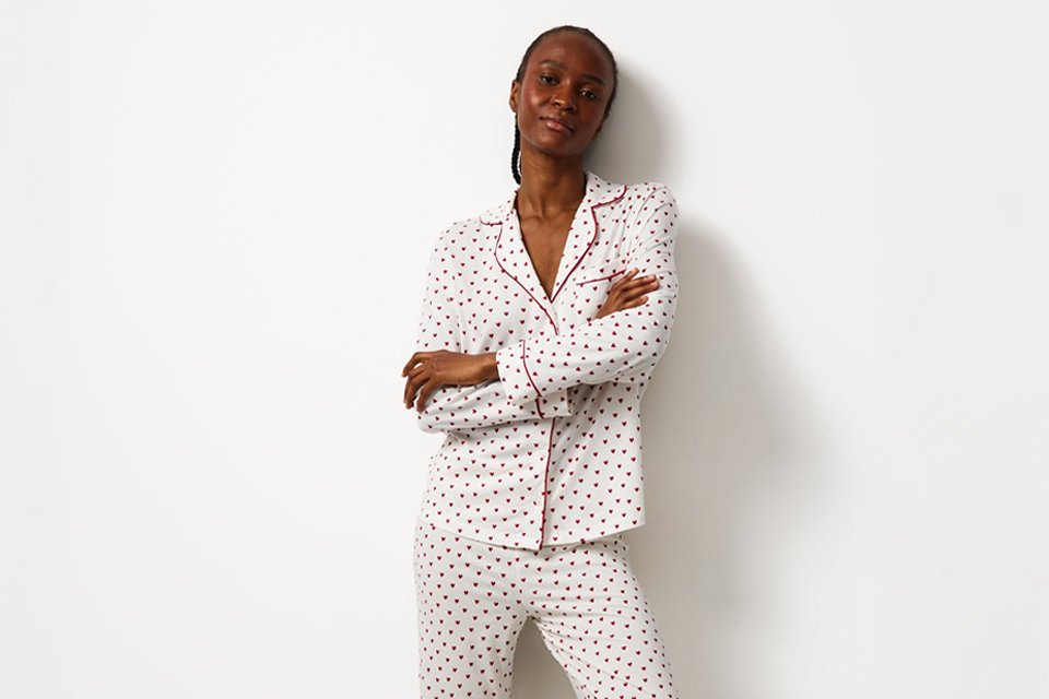 Person standing against a white wall wearing a white pajama set with small black and red heart prints, red piping detail, and beige fluffy slippers.